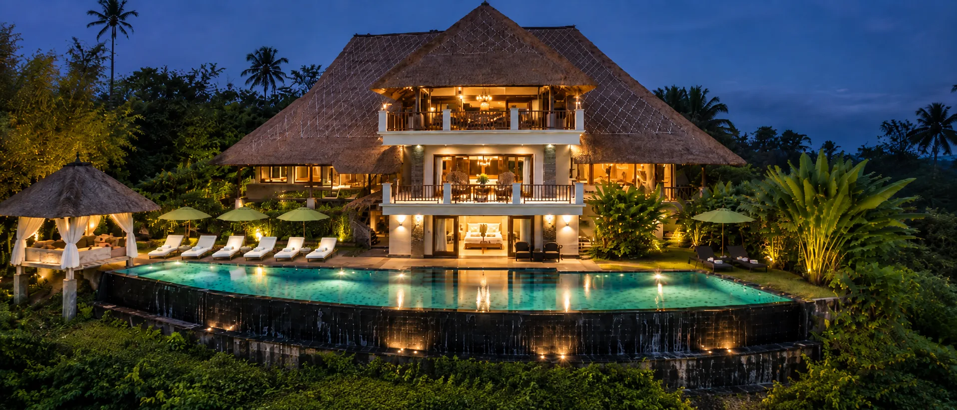 Illuminated thatched-roof building and curved swimming pool beneath a deep blue evening sky