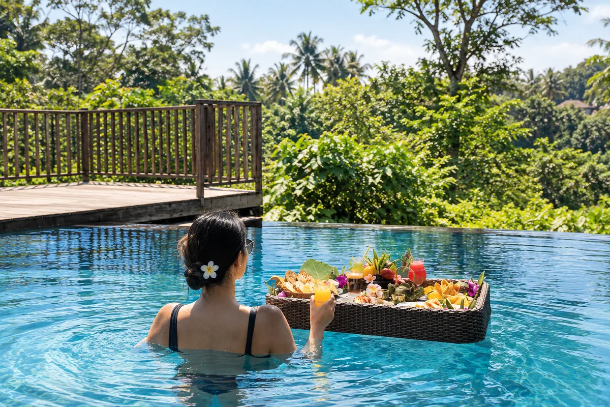 Guest in a swimming pool beside a floating tray of food, with a timber railing and tropical trees beyond