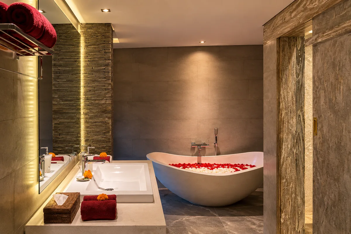 White freestanding bathtub with red petals beside a washbasin and softly illuminated mirror