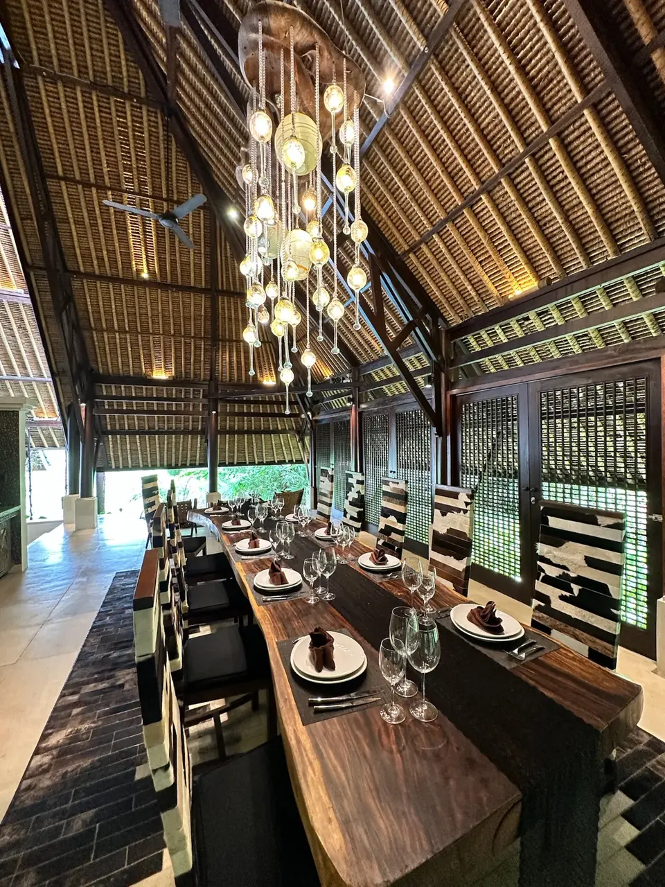 Long wooden dining table set beneath the estate’s open-air thatched pavilion, with hanging lights and garden views