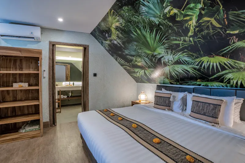 Green Room 2 bed beside a timber storage cabinet and bathroom entrance beneath a tropical green mural