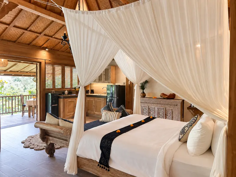 Large double bed in Main Joglo Room beneath white canopy drapes, with carved cabinets and warm timber details