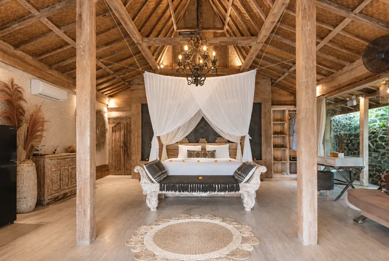 Main Joglo Room with a draped double bed, soaring woven timber roof, chandelier, carved bench and sitting area
