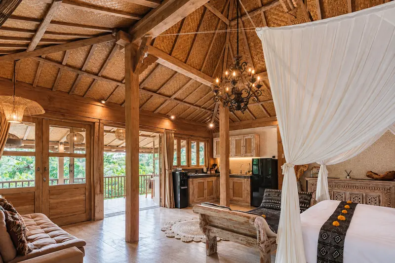 Wide view across Main Joglo Room showing the canopy bed, sitting area, kitchenette, timber ceiling and balcony doors