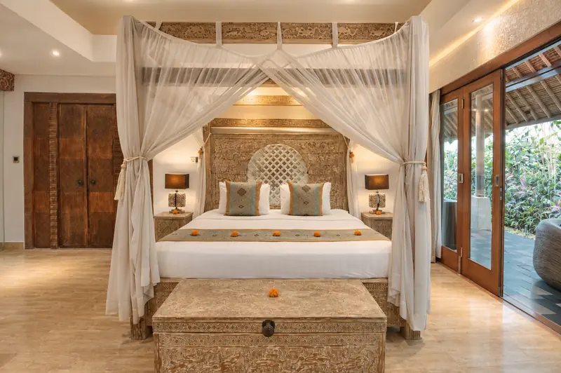 Red Room bedroom with a carved canopy bed, white drapes, timber wardrobe and tall glazed doors opening to tropical greenery