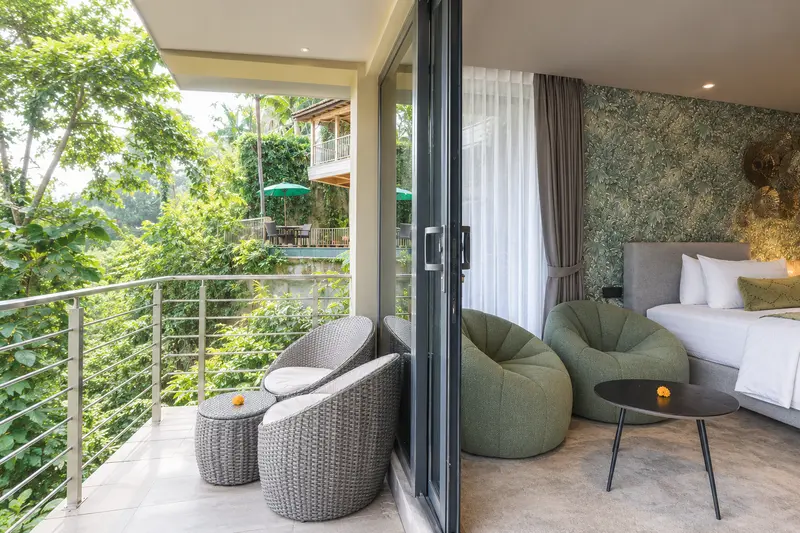 River Villa Room 1 balcony seating beside glass doors, with green lounge chairs and a twin bed inside