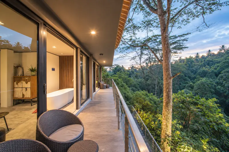 Long furnished balcony outside River Villa Room 1 overlooking dense tropical treetops at dusk