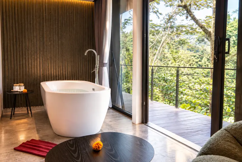 Freestanding bathtub in River Villa Room 1 beside full-height glass doors and a tropical view
