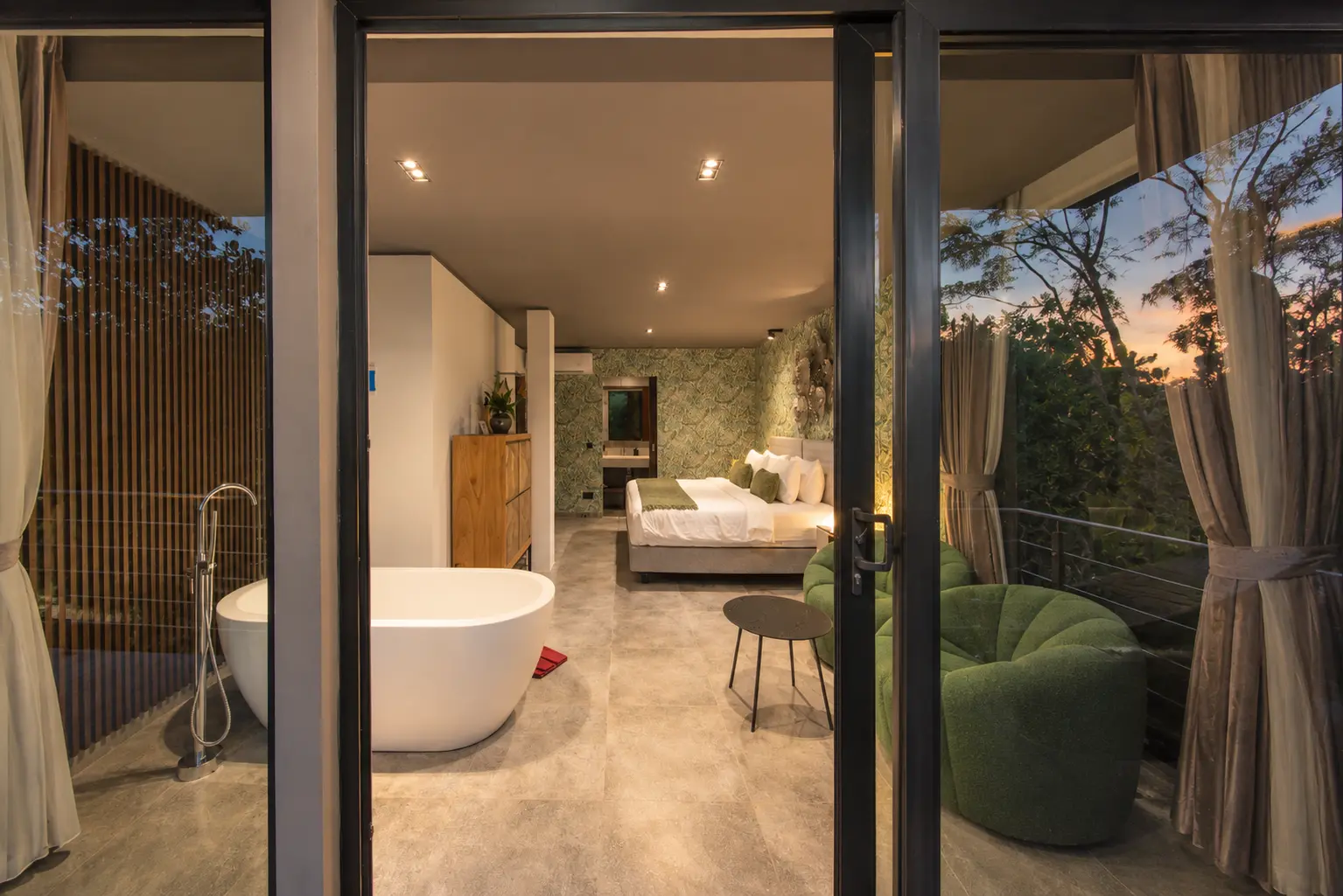 River Villa Room 2 at dusk with a freestanding bath, glazed doors and a warmly lit bedroom beyond