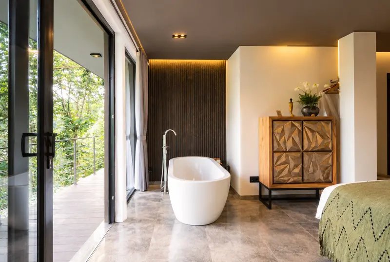 Freestanding bathtub in River Villa Room 2 beside full-height glass doors and a carved cabinet