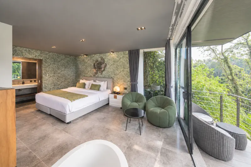 River Villa Room 2 seen across the bathtub towards twin beds, lounge seating, balcony chairs and tropical foliage