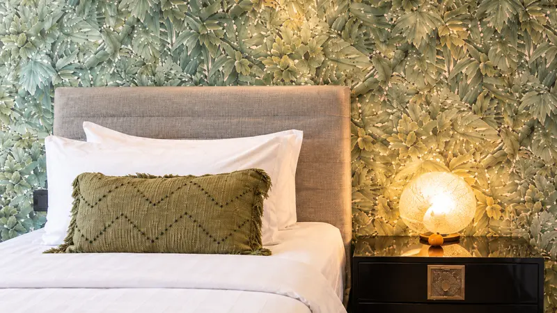 Close view of a River Villa Room 3 single bed with woven olive cushion, botanical wall and glowing lamp