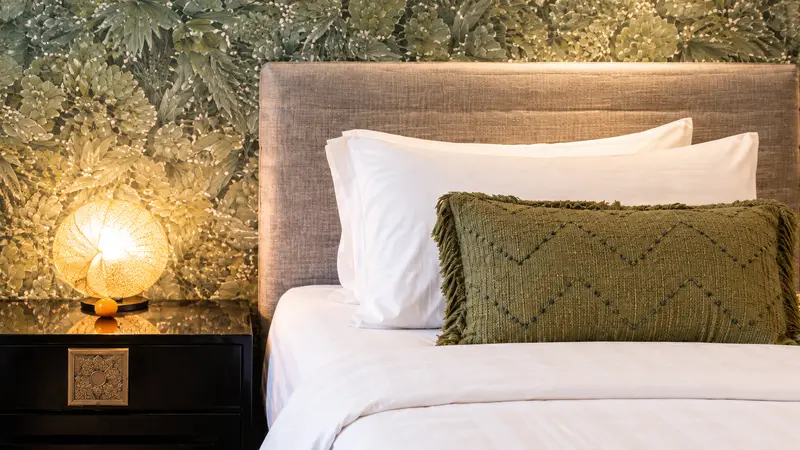 Close view of River Villa Room 4 bed linen, woven olive cushion, bedside lamp and botanical wall covering