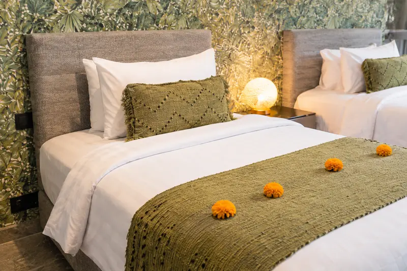 Close view of a River Villa Room 5 single bed with olive woven cover, orange flowers and glowing bedside lamp
