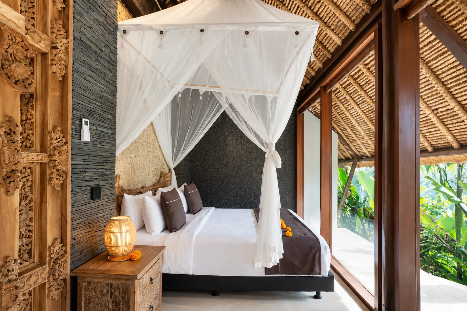 Studio Room 2 with ornate carved timber, a draped bed, exposed roof and tropical planting beyond glazed doors
