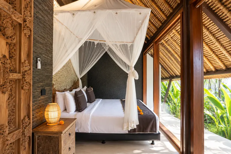 Side view of Studio Room 2 with canopy bed, carved doorway and tropical leaves beyond full-height glazing